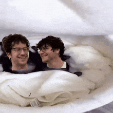 two men are laying on a bed with a white blanket
