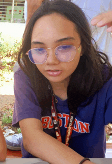 a girl wearing glasses and a boston shirt is sitting at a table