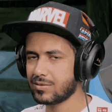 a man wearing a marvel hat and headphones looks at the camera