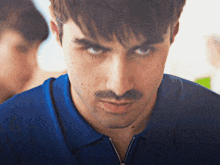 a man with a mustache wearing a blue shirt looks at the camera