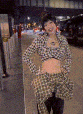 a woman in a plaid shirt is standing on a sidewalk with her hands on her hips and smiling .