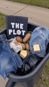 a book titled the plot sits on top of a wheelbarrow filled with clothes and potatoes