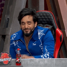 a man in a blue jacket is sitting at a table with a bottle of charged soda