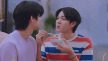 two young men are sitting next to each other and one of them is feeding the other a bowl of food .