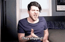 a man in a blue shirt is sitting on a couch and saying `` they shipped ! ''
