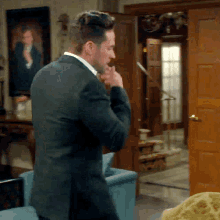 a man in a suit is standing in a living room with his hand on his chin