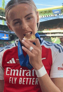 a woman wearing an emirates fly better jersey holds a medal in her mouth