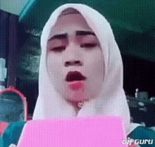 a woman wearing a white hijab is holding a pink piece of paper .