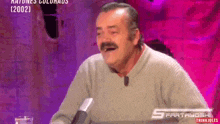 a man with a mustache is laughing while sitting in front of a microphone .