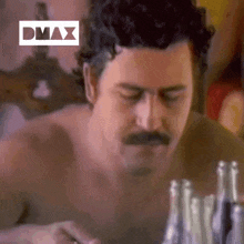 a shirtless man with a mustache is sitting at a table with many bottles of alcohol .