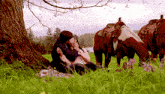 a couple sitting under a tree in a field with horses in the background