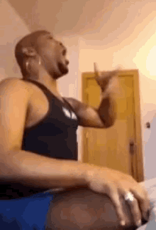 a man in a black tank top is sitting on a bed and drinking from a cup .