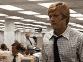 a man in a plaid shirt and tie stands in a busy office