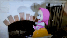 a doll in a pink head scarf is sitting on a yellow object in front of a fireplace