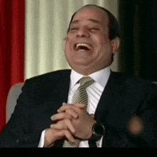 a man in a suit and tie is laughing while sitting in a chair with his hands folded .