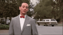 a man in a suit and bow tie is standing in front of a car on the street .