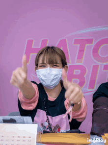 a woman wearing a mask giving a thumbs up in front of a pink background that says hatc bit