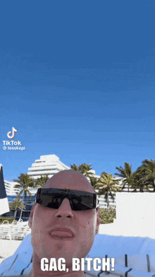 a bald man wearing sunglasses is laying on a beach and says " gag bitch "