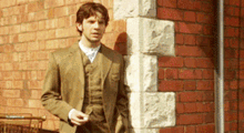 a man in a suit leans against a brick wall holding a cigarette