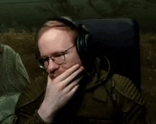 a man wearing headphones and glasses is sitting in a chair covering his mouth with his hand .