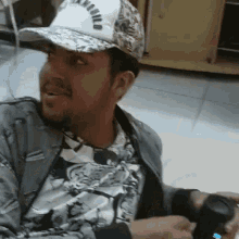 a man wearing a hat and a jacket is sitting on the floor holding a remote control .