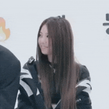 a woman with long brown hair is wearing a black jacket and a necklace while standing next to a man .