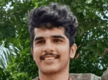 a young man with a mustache is smiling for the camera in front of trees .