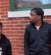 two men are standing in front of a brick wall and one of them is wearing a hat .
