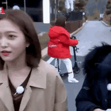 a woman in a trench coat is standing next to a person riding a scooter on a street .