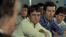 a group of men are sitting at a table with their arms crossed and one man is looking at the camera