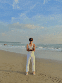 a man in white pants and flip flops stands on a beach with his arms crossed