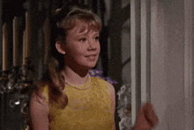 a young girl in a yellow dress and pearls is standing in front of a window .