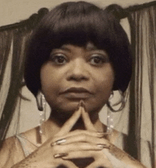 the woman is wearing a wig and earrings and has her hands folded in prayer .