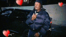 a man in a black jacket is sitting in front of a car with hearts flying around him .