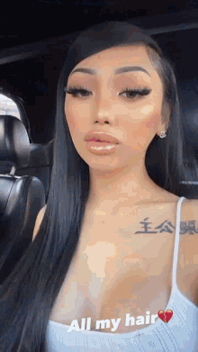 a woman with long black hair is sitting in a car wearing a white tank top and a tattoo on her chest .