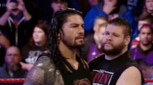 roman reigns is standing next to a man in a wrestling ring in front of a crowd .