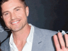 a man in a suit and white shirt is smiling and holding up his hand