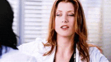 a woman in a white lab coat is sitting in front of a window and making a funny face .