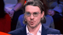 a man wearing glasses and a suit is talking into a microphone while sitting in front of a crowd .