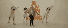 a man in a suit and tie stands in front of a group of women dancing in front of the letters b and n