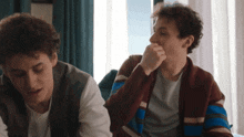 two young men are sitting next to each other and one has his hand on his nose