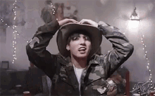 a young man wearing a cowboy hat and a camouflage jacket is making a funny face .