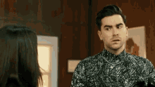 a man in a black and white shirt is talking to a woman in a room and saying `` fine '' .