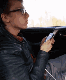 a young man wearing glasses is sitting in a car looking at his cell phone