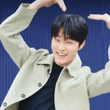 a young man in a green jacket is smiling and making a heart with his hands