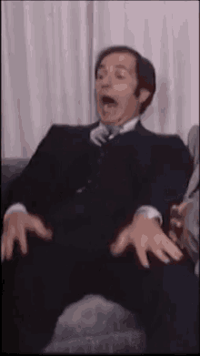 a man in a suit and tie is sitting on a couch with his mouth open and his hands outstretched .