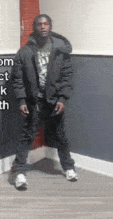 a man in a black jacket is standing in the corner of a room smoking a cigarette .