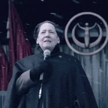 a woman in a black cape is holding a microphone in front of a circle with the letter g on it