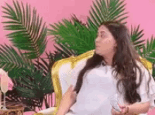 a woman is sitting in a chair with a pink background and palm leaves behind her .