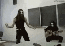 a man with long hair is dancing in front of a blackboard in a classroom while another man plays a guitar .